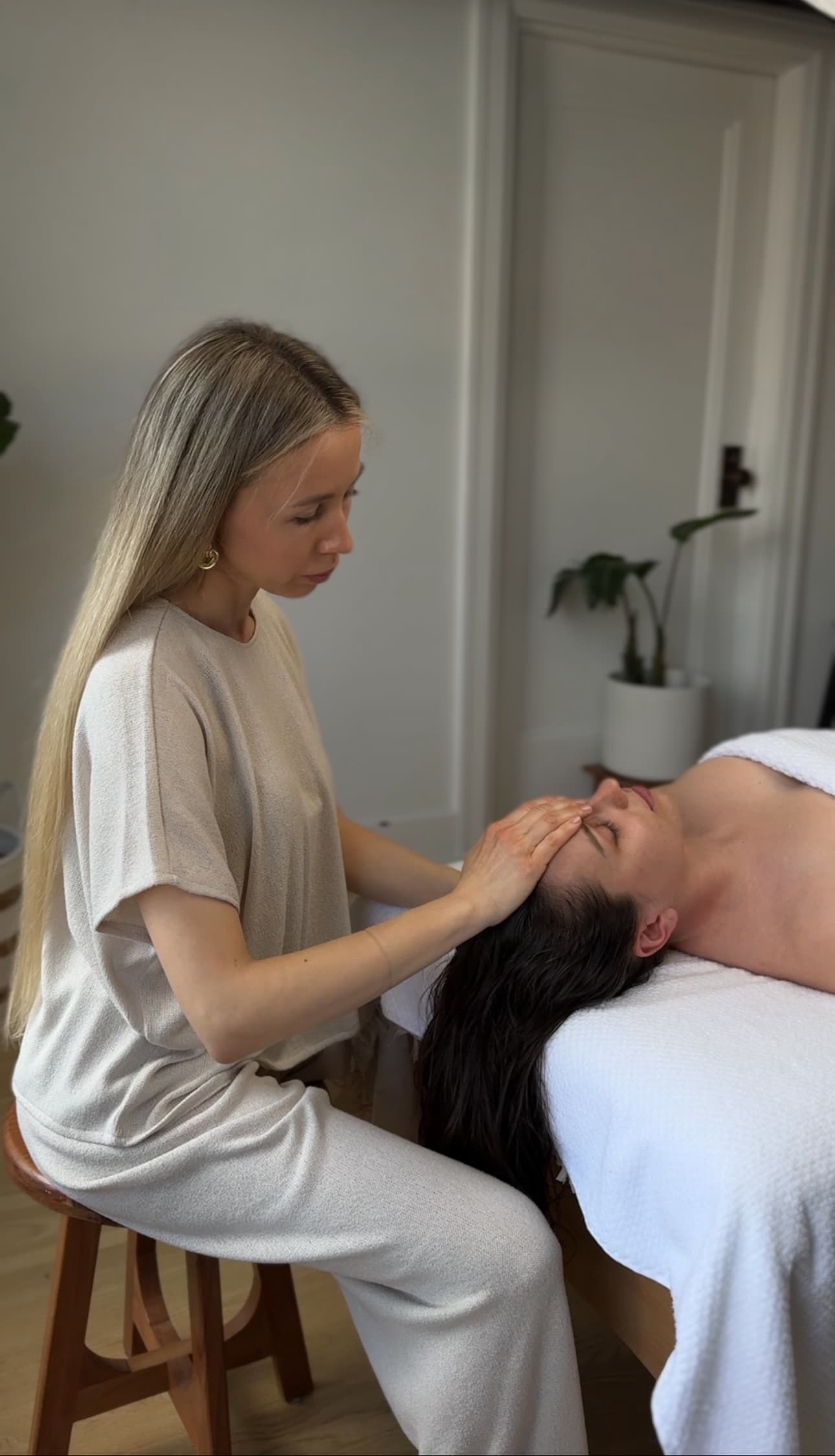 Blonde woman performs a facial massage on a client lying on a white massage table.