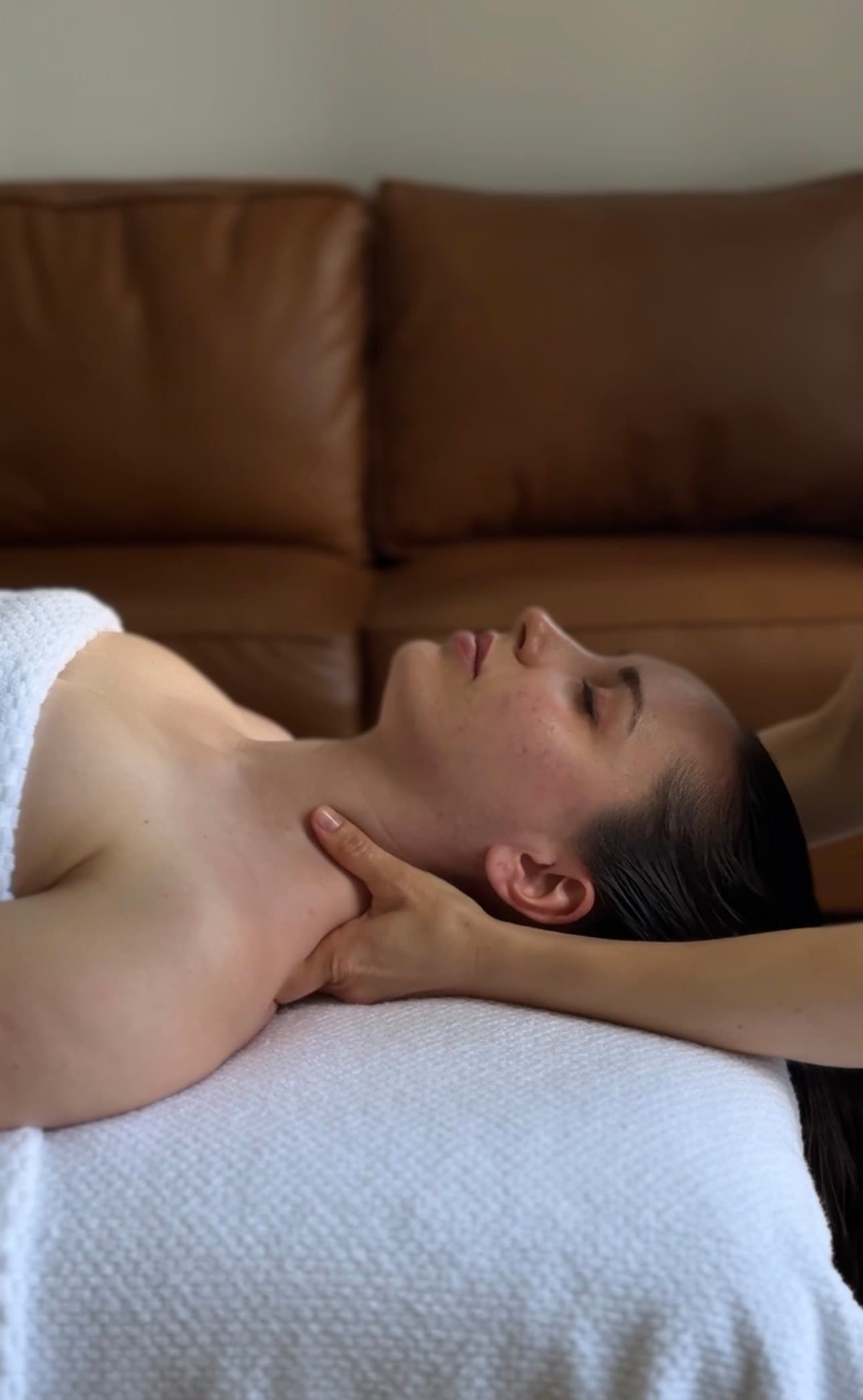Woman lying on a white towel receiving a relaxing neck massage with eyes closed.